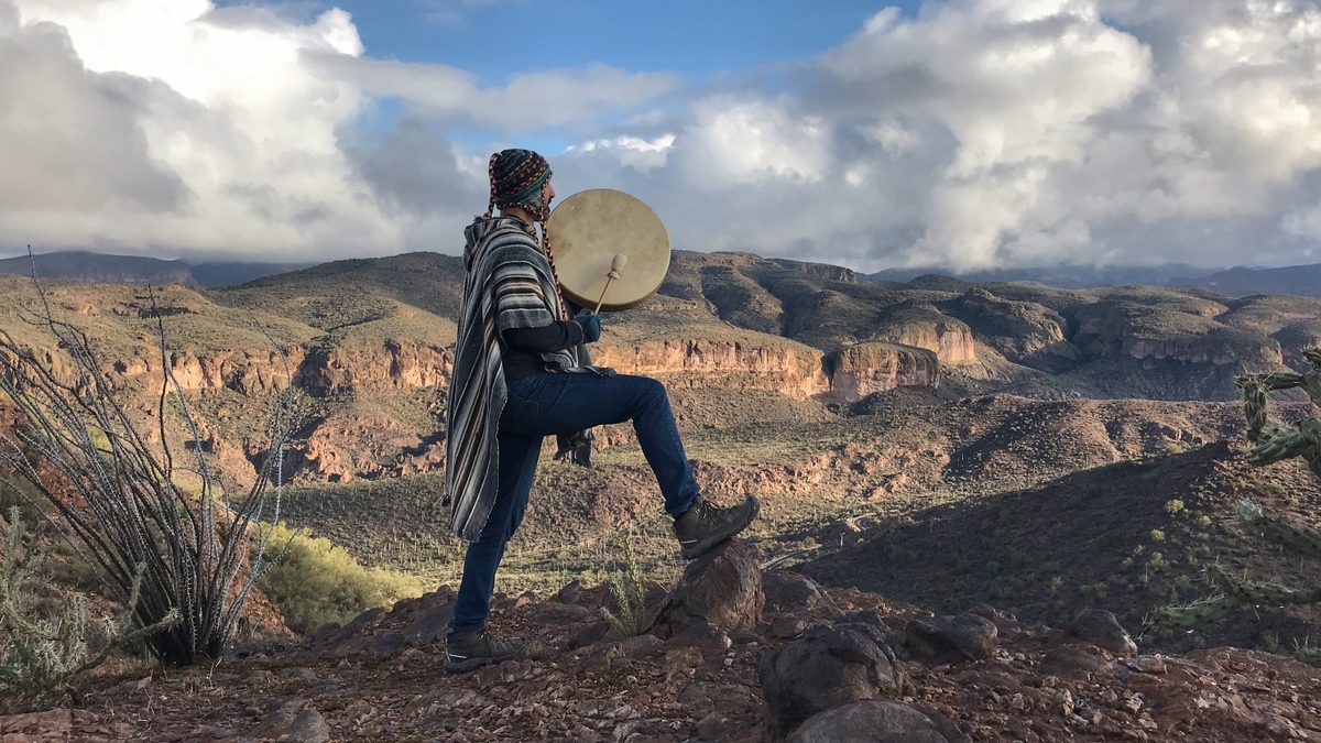 Someone in a striped poncho with a hand drum, one foot up on a rock at the rim of a wide desert canyon in afternoon light