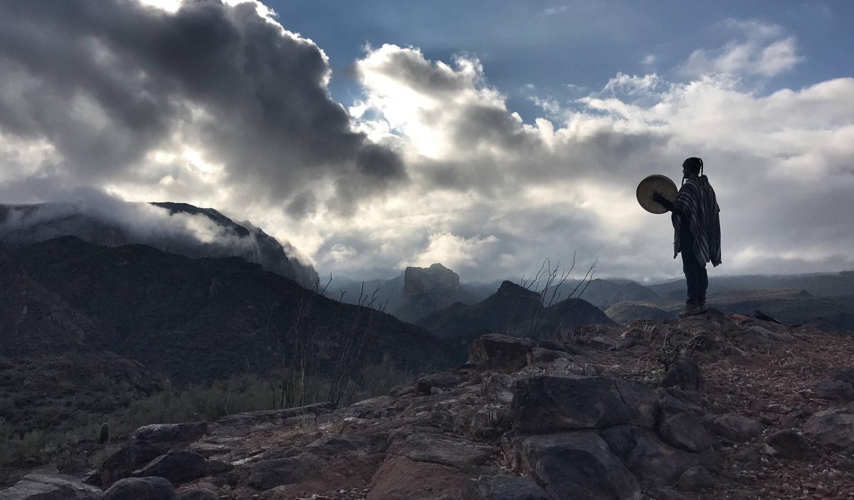 A figure alone on a rock ridge at dusk holding a round hand drum up against a break in a storm cloud, desert mountains below