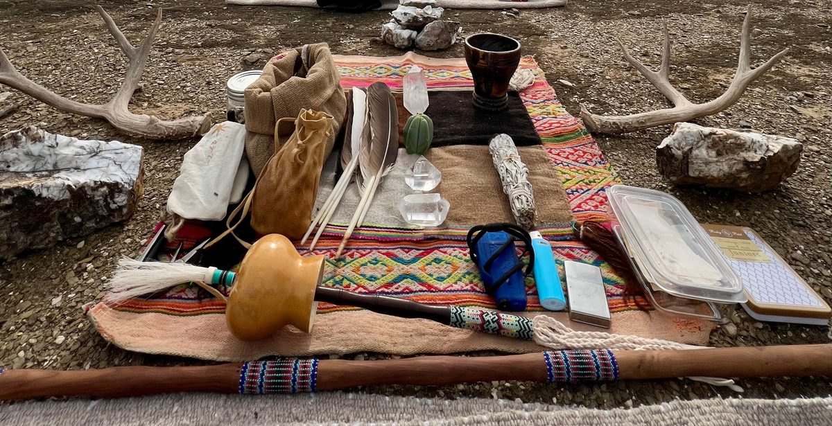 A mesa laid out on gravel ground in Arizona: a woven Andean cloth holding a gourd rattle trailing white horse hair, feathers, crystals, a small green cactus at the center, a bundle of sage and a dark cup, with a beaded staff along its front edge, antlers to either side and white stones beside them