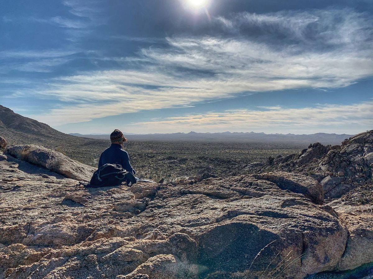 Someone sitting on bare rock at the edge of a high place, looking out over a wide desert basin under a long band of cloud