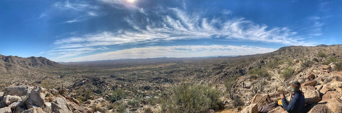 A wide view along a desert basin from high ground, ridges running away on both sides under a long sky, one walker small on the rocks