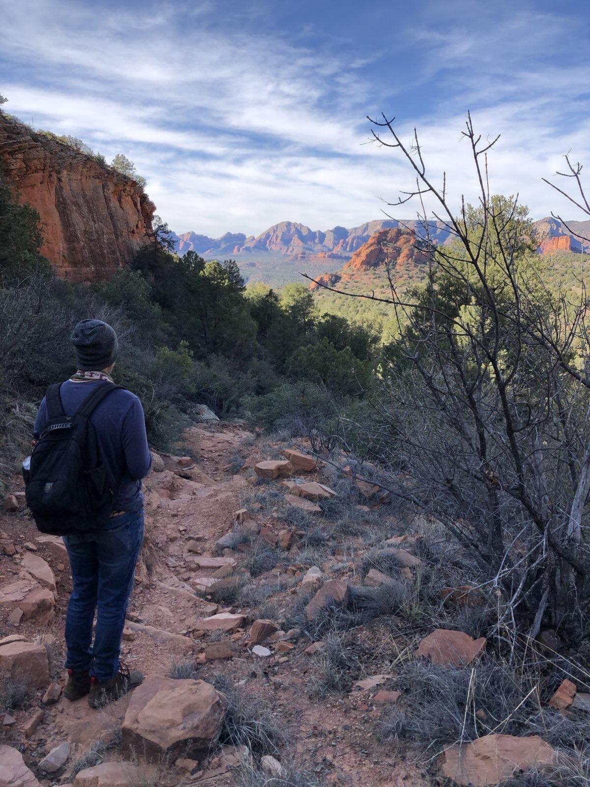 A walker on a rocky trail through juniper scrub with red sandstone cliffs standing beyond