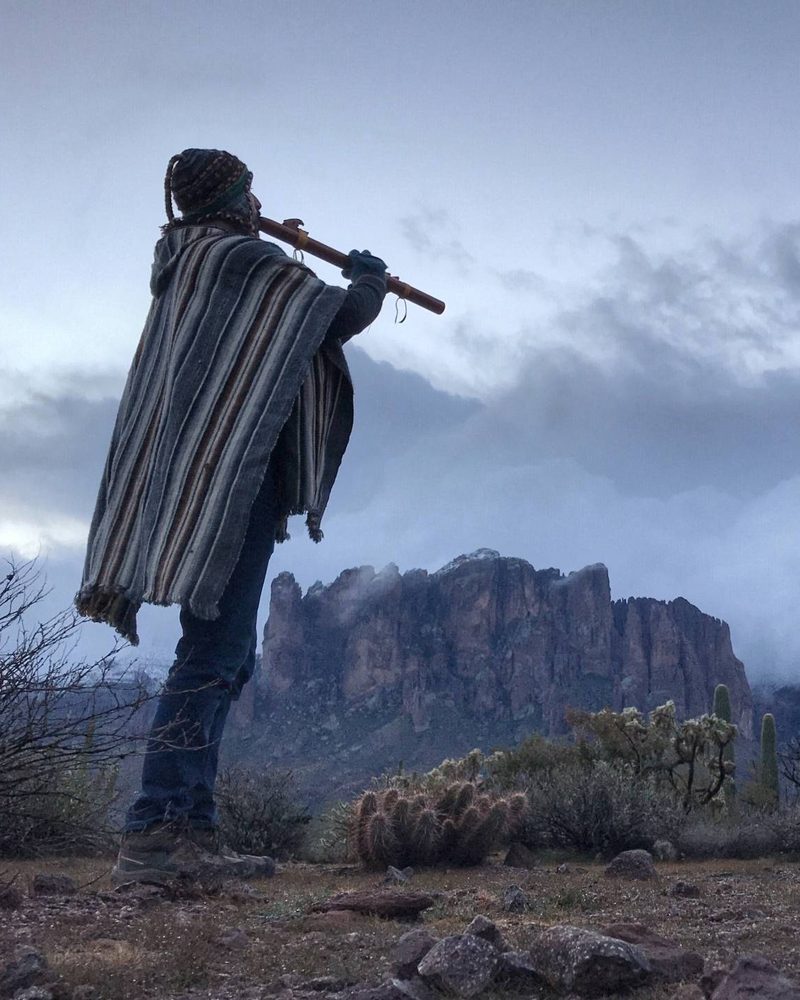 A walker in a striped poncho playing a long wooden flute, the Superstition Mountains standing in mist behind