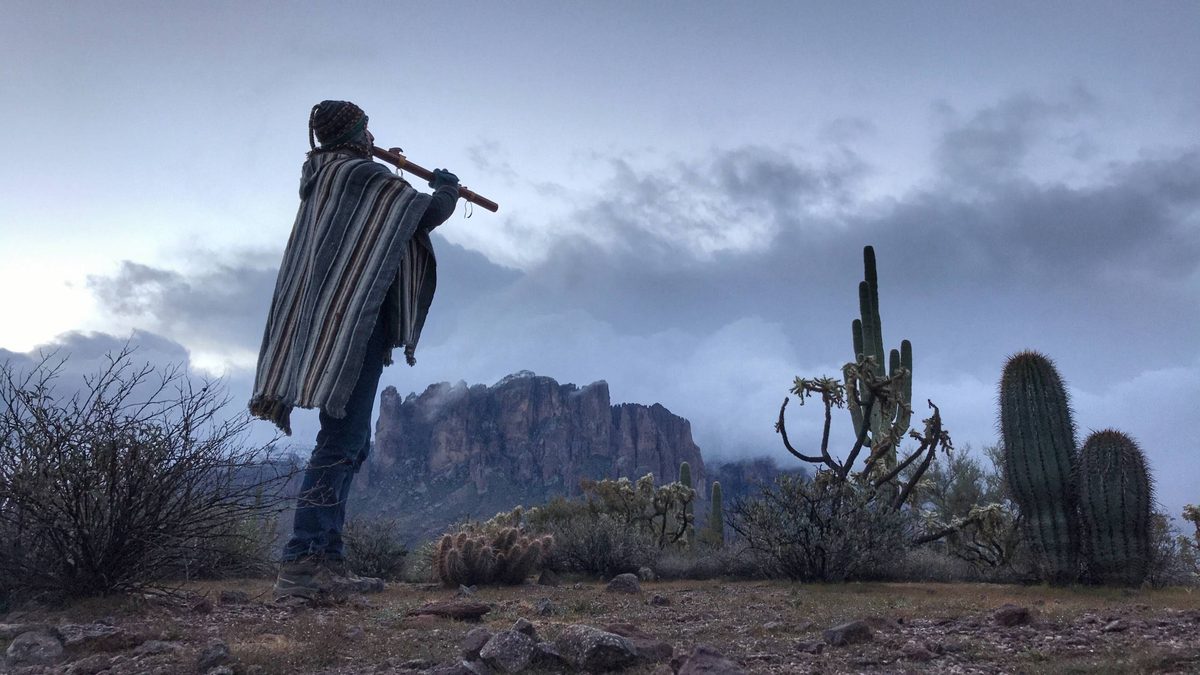 A walker in a striped poncho playing a long wooden flute among saguaro, with the Superstition Mountains standing in mist behind